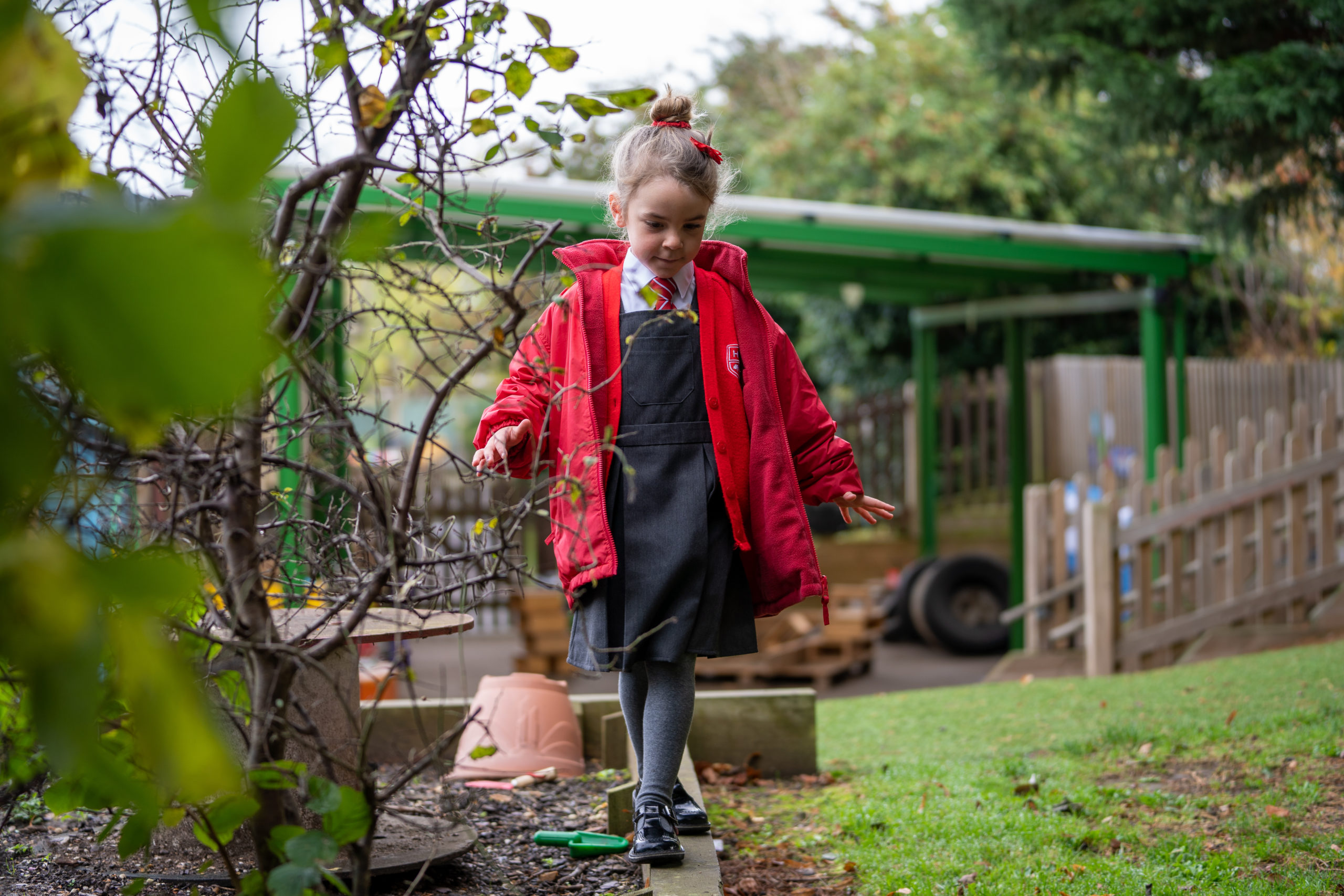 A young girl is pictured walking along a wooden plank outside on the academy grounds. She is wearing her winter coat, as it is cloudy outside.