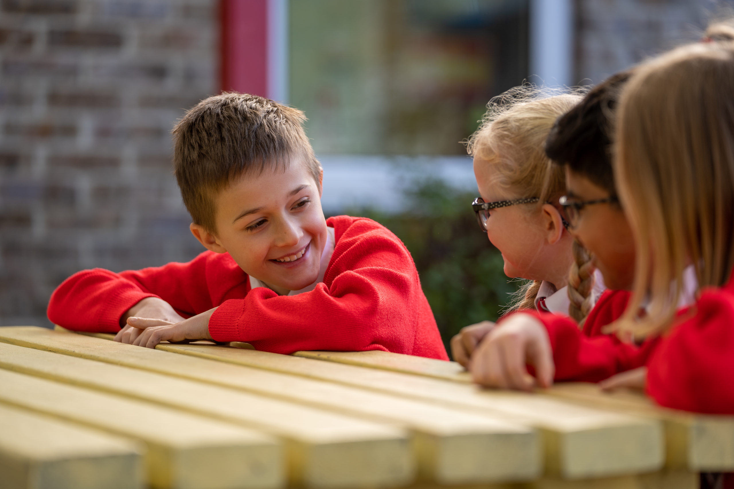 A group of four young pupils, two boys and two girls, are pictured sat together at a table outdoors on the academy grounds, smiling at one another.