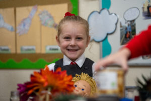 A young girl is pictured smiling for the camera in her academy uniform, whilst playing with a doll in her arms.